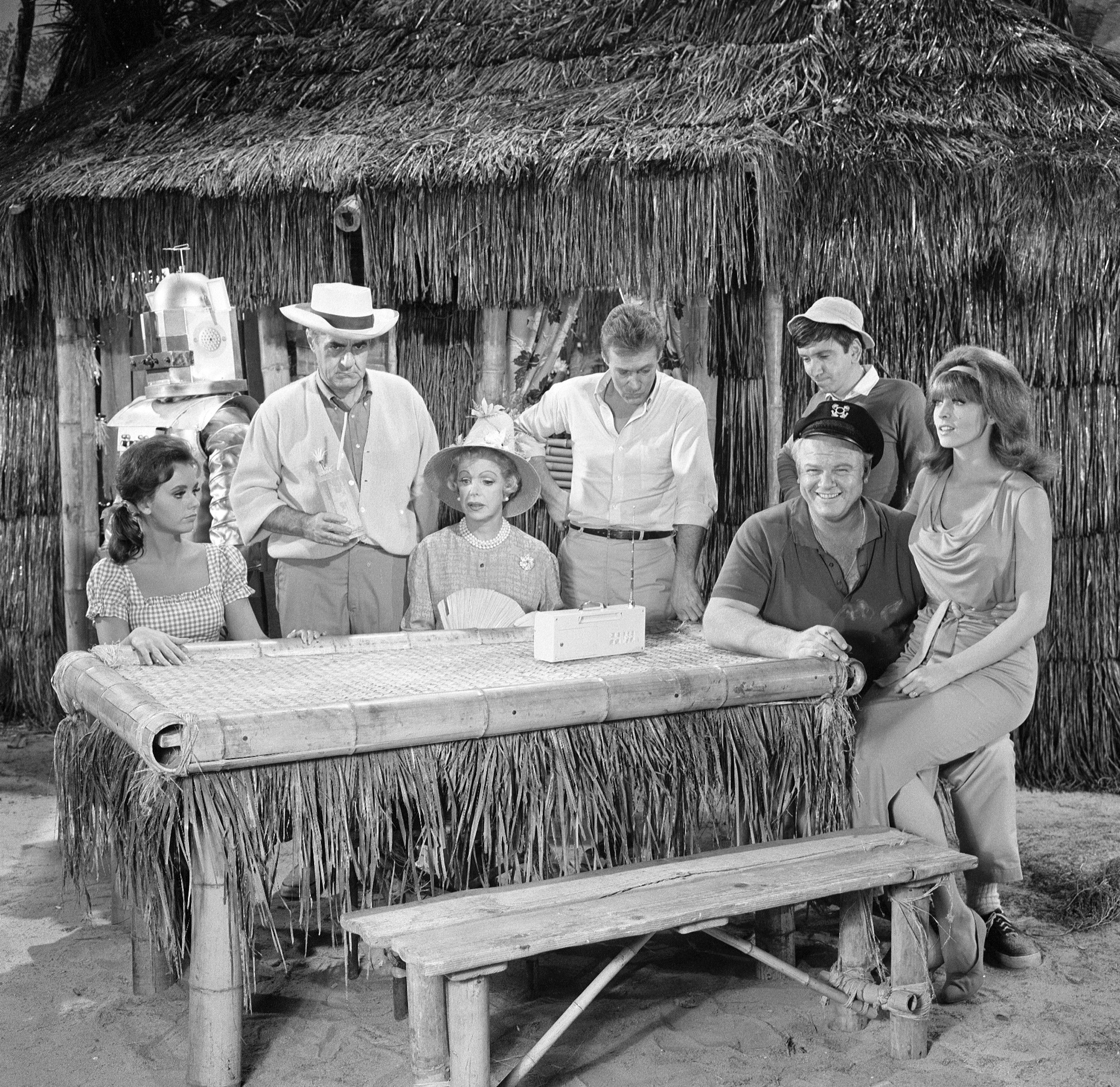 'The Gilligan's Island' cast sitting around a picnic table near a bamboo hut.