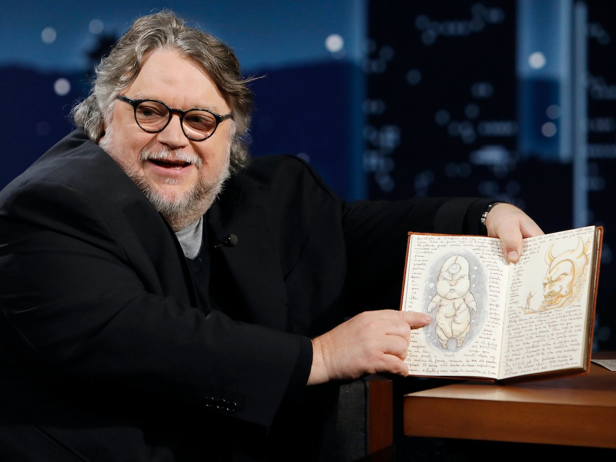Guillermo del Toro wears dark glasses and a dark coat as he points to an illustration o a creature in a book on ‘Jimmy Kimmel Live!’