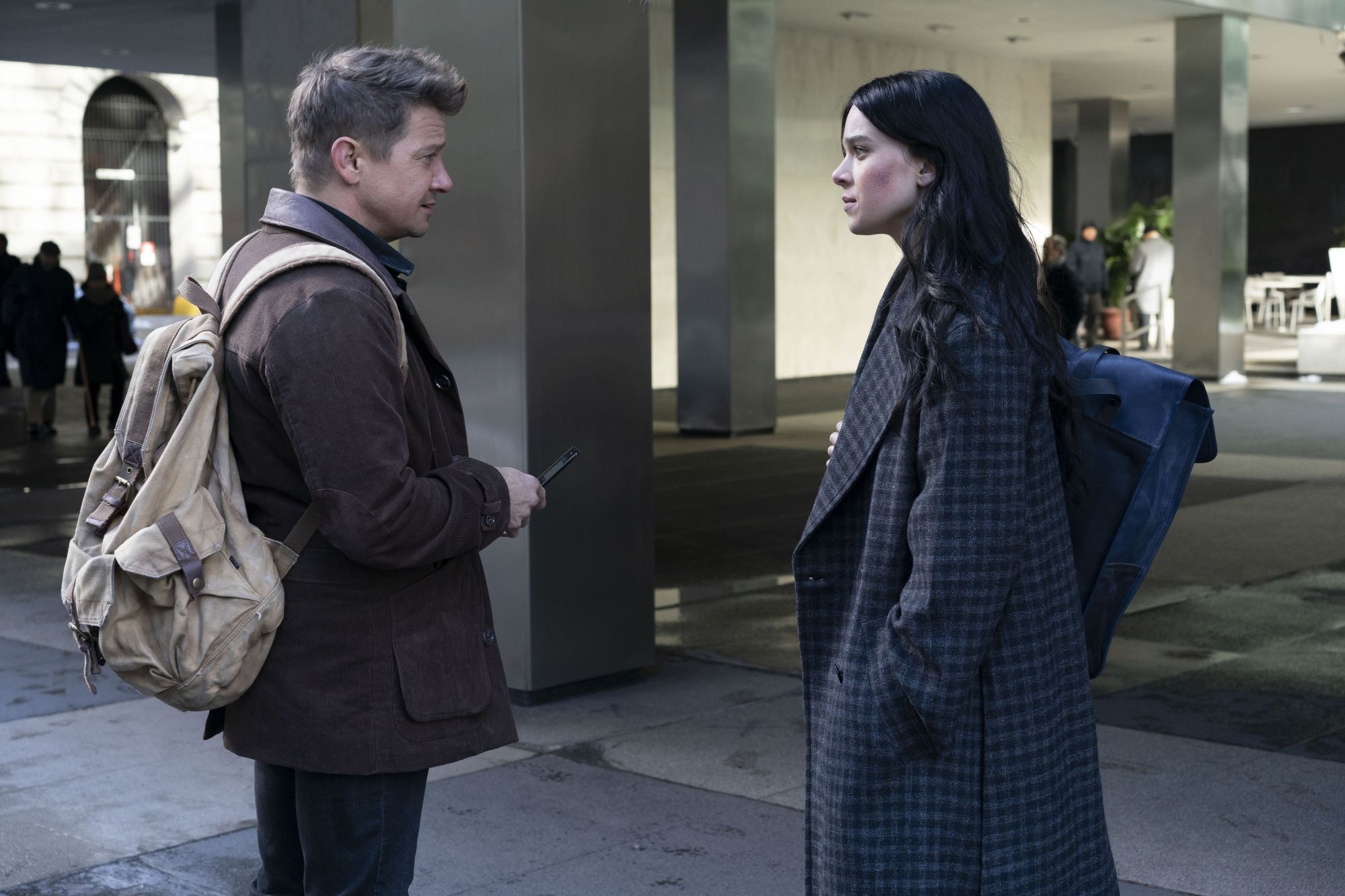 'Hawkeye' Episode 4 stars Jeremy Renner and Hailee Steinfeld, in character as Clint Barton and Kate Bishop, stand outside of a building. Clint wears a brown coat over black pants and has a tan backpack slung over his shoulders. Kate wears a dark gray plaid coat and has a black bag slung over her shoulder. Renner also starred in 'Avengers: Endgame.'