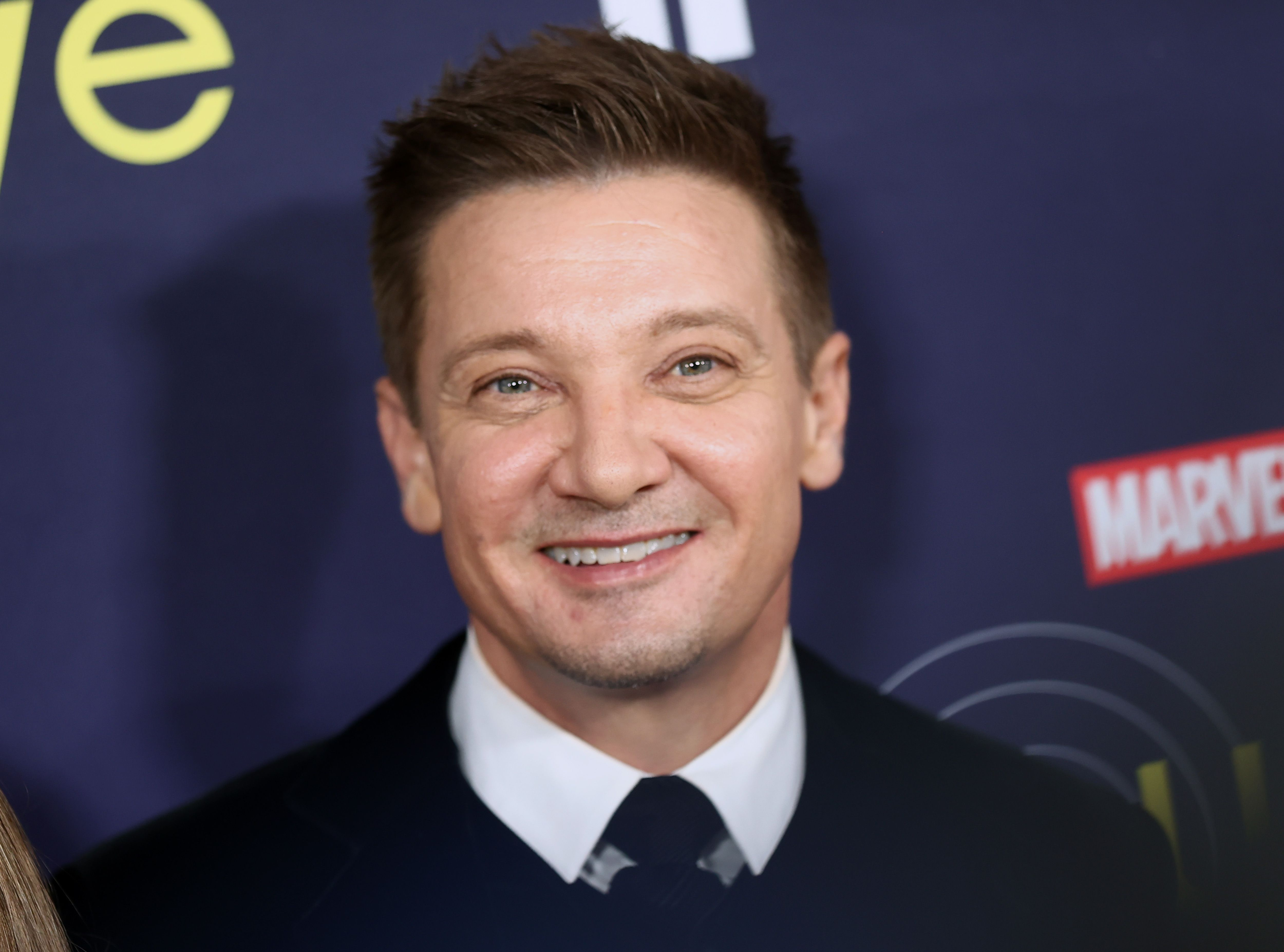 'Hawkeye' star Jeremy Renner wearing a white collared shirt and black suit. He's smiling at the camera and standing in front of a wall with the Disney+ show's logo.