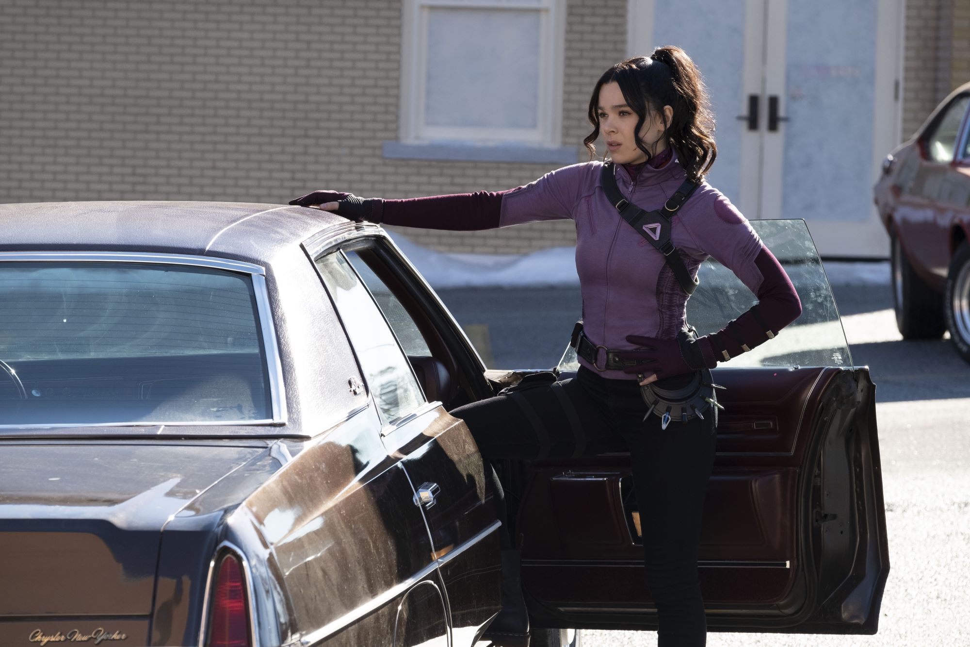 'Hawkeye' star Hailee Steinfeld as Kate Bishop, who found a mysterious rolex watch in episode 4, stands beside the passengers seat of a car and wears her superhero suit: a purple long-sleeved shirt and black pants.