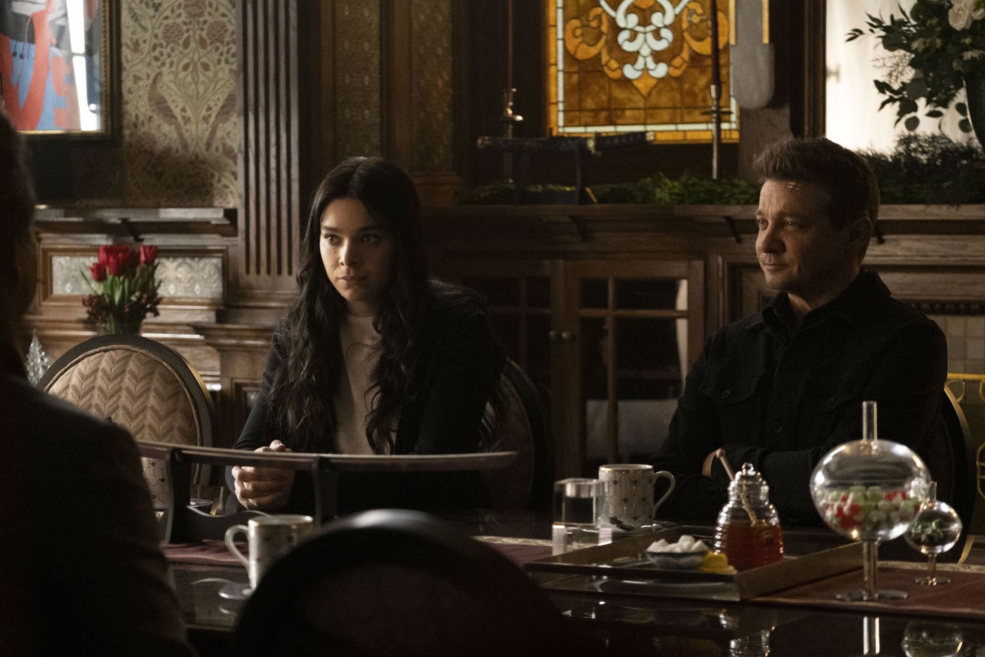 'Hawkeye' Episode 5 stars Hailee Steinfeld and Jeremy Renner, in character as Kate Bishop and Clint Barton, sit at a table. Kate wears a black jacket over a white shirt. Clint wears a black button-up jacket.