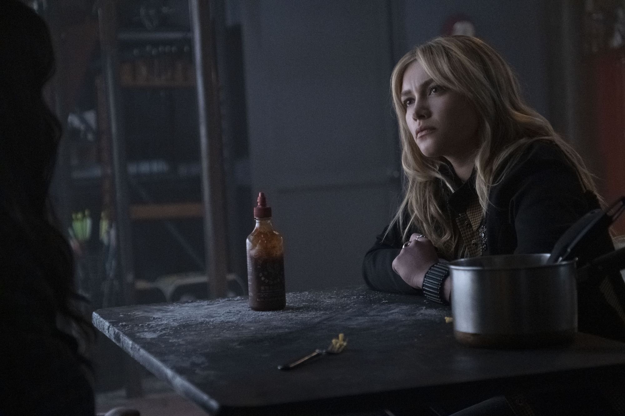 'Hawkeye' star Florence Pugh, in character as Yelena Belova, acts opposite of Hailee Steinfeld's character Kate Bishop in episode 5 in the mac and cheese scene. Yelena wears a black and brown jacket and sits at a table with a pot of mac and cheese and a bottle of hot sauce on it.