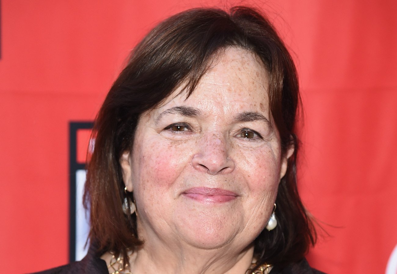 Ina Garten smiles wearing a black top while standing in front of a red background