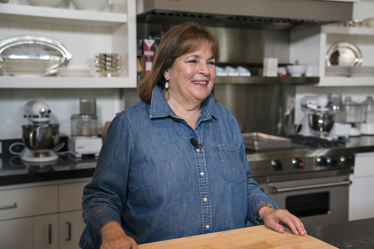 Ina Garten smiles as she stands at a butcher block wearing a blue button down shirt