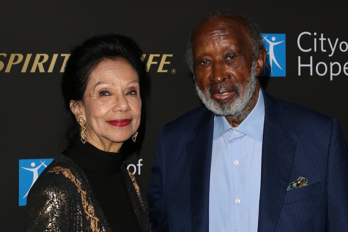 Jacqueline Avant, wearing a black and gold jacket, poses next to Clarence Avant wearing a blue suit.