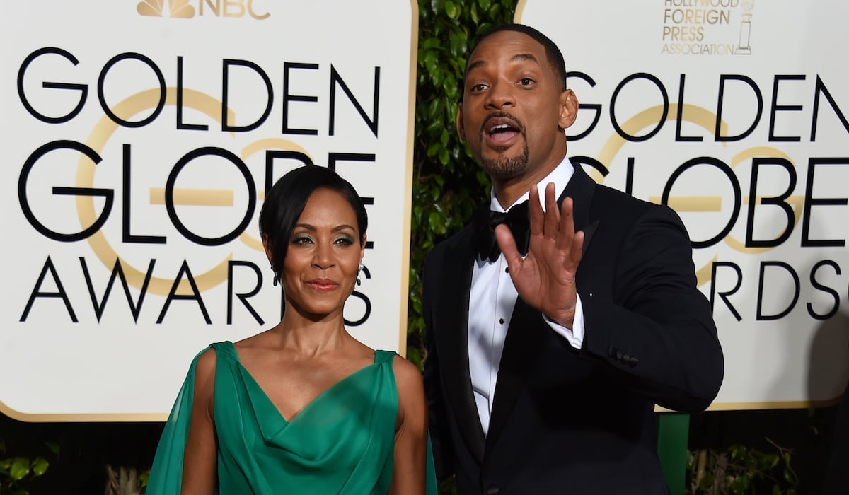 Jada Pinkett Smith wears a green dress and Will Smith wears a suit and holds his hand up while speaking on the red carpet at the 2016 Golden Globes