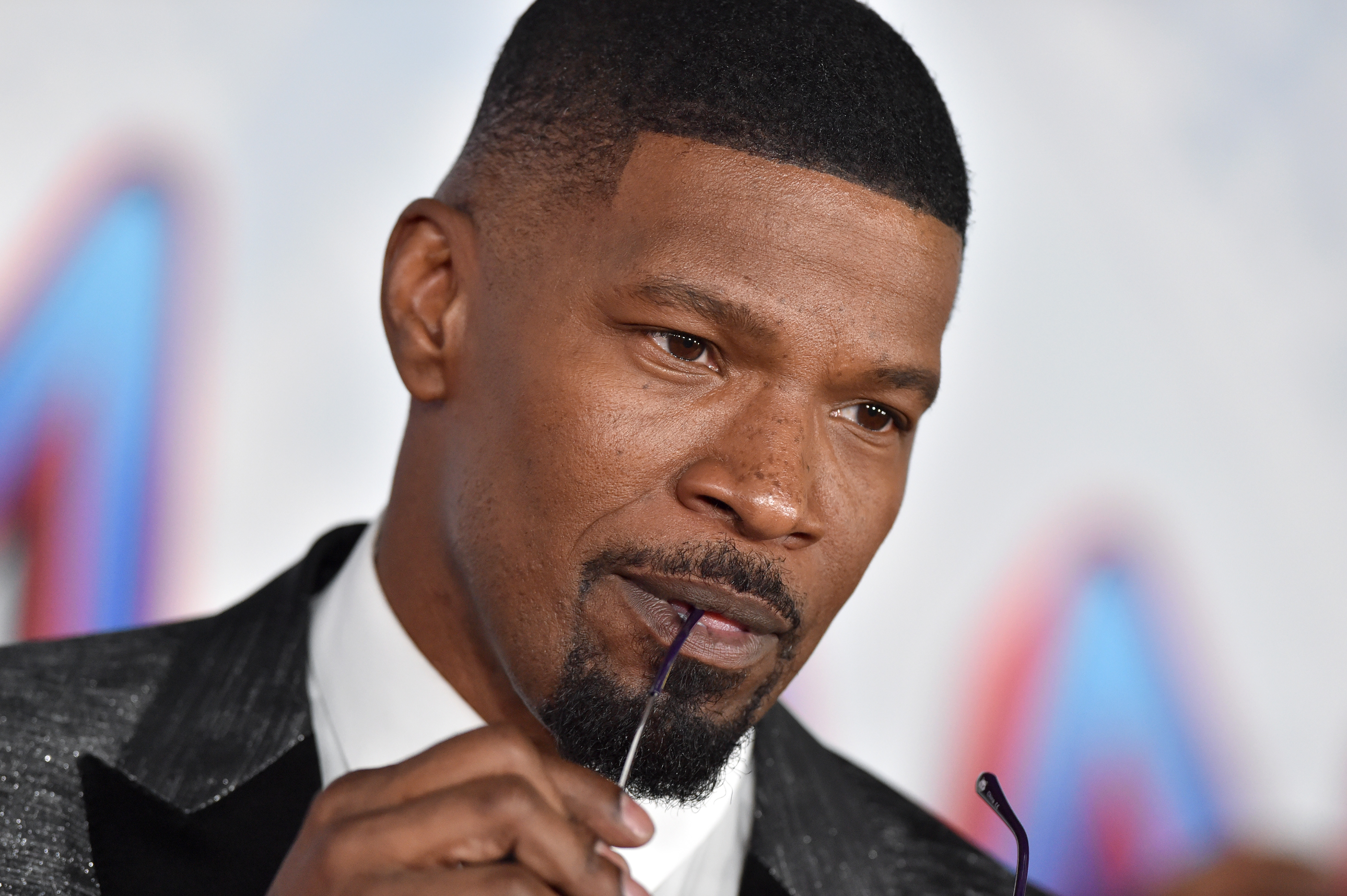 'Spider-Man: No Way Home' star Jamie Foxx wears a silver sparkly suit over a white button-up shirt and bites the end of his glasses that he's holding in his hand.