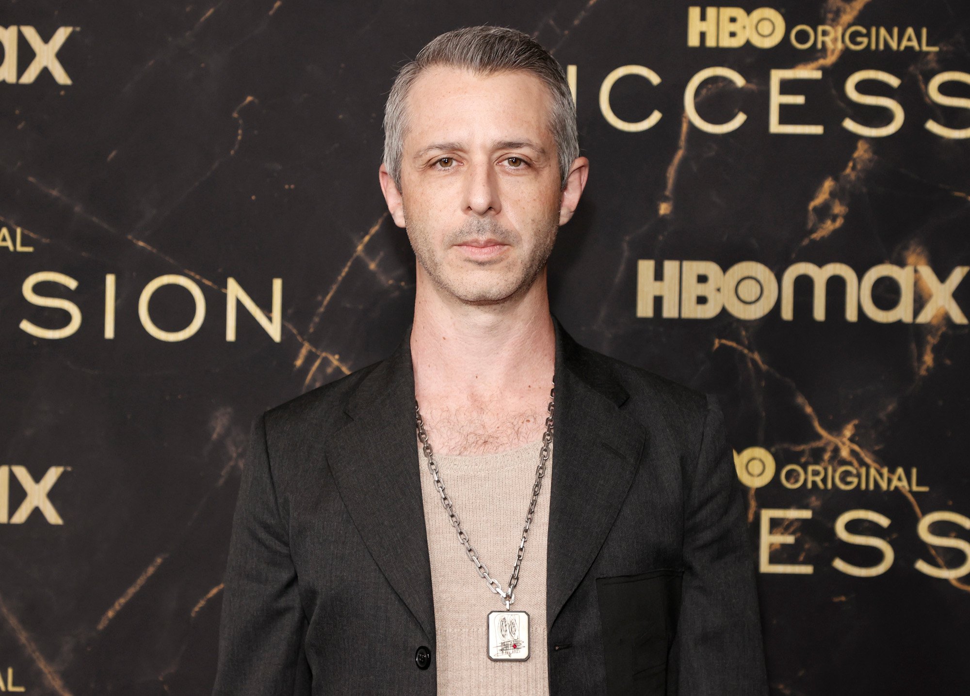 'Succession' star Jeremy Strong a tan shirt, black jacket, and gold chain. He's standing in front of a wall that says 'HBO Max' and 'Succession,' and he's looking directly at the camera.