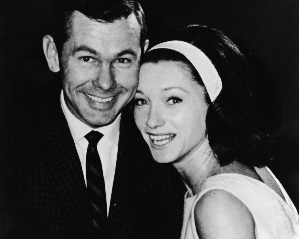 Johnny Carson in a suit and tie, smiles next to his second wife, Joanne, in a dress and matching headband