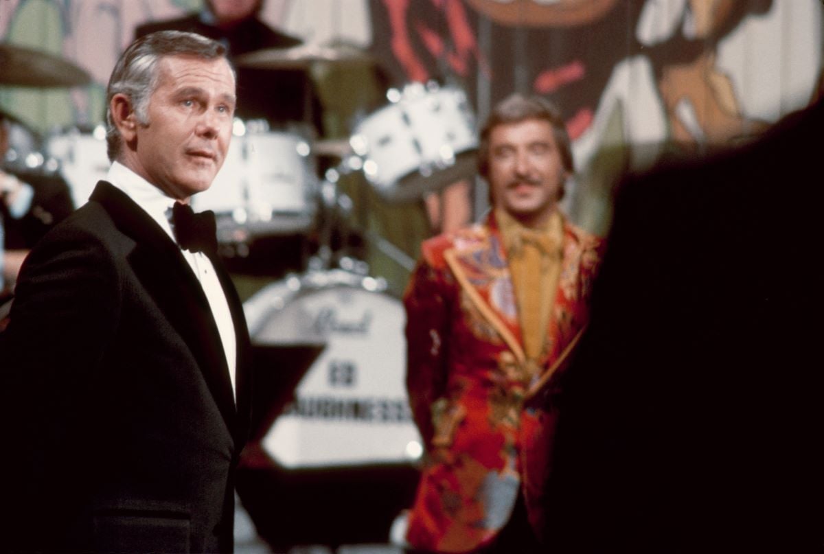 Johnny Carson stands in front of the 'Tonight Show' band, wearing a black and white suit and bow-tie