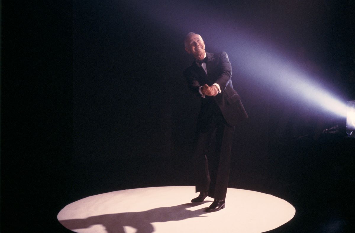 Johnny Carson in a black suit and tie, simulating a golf swing while standing in a spotlight