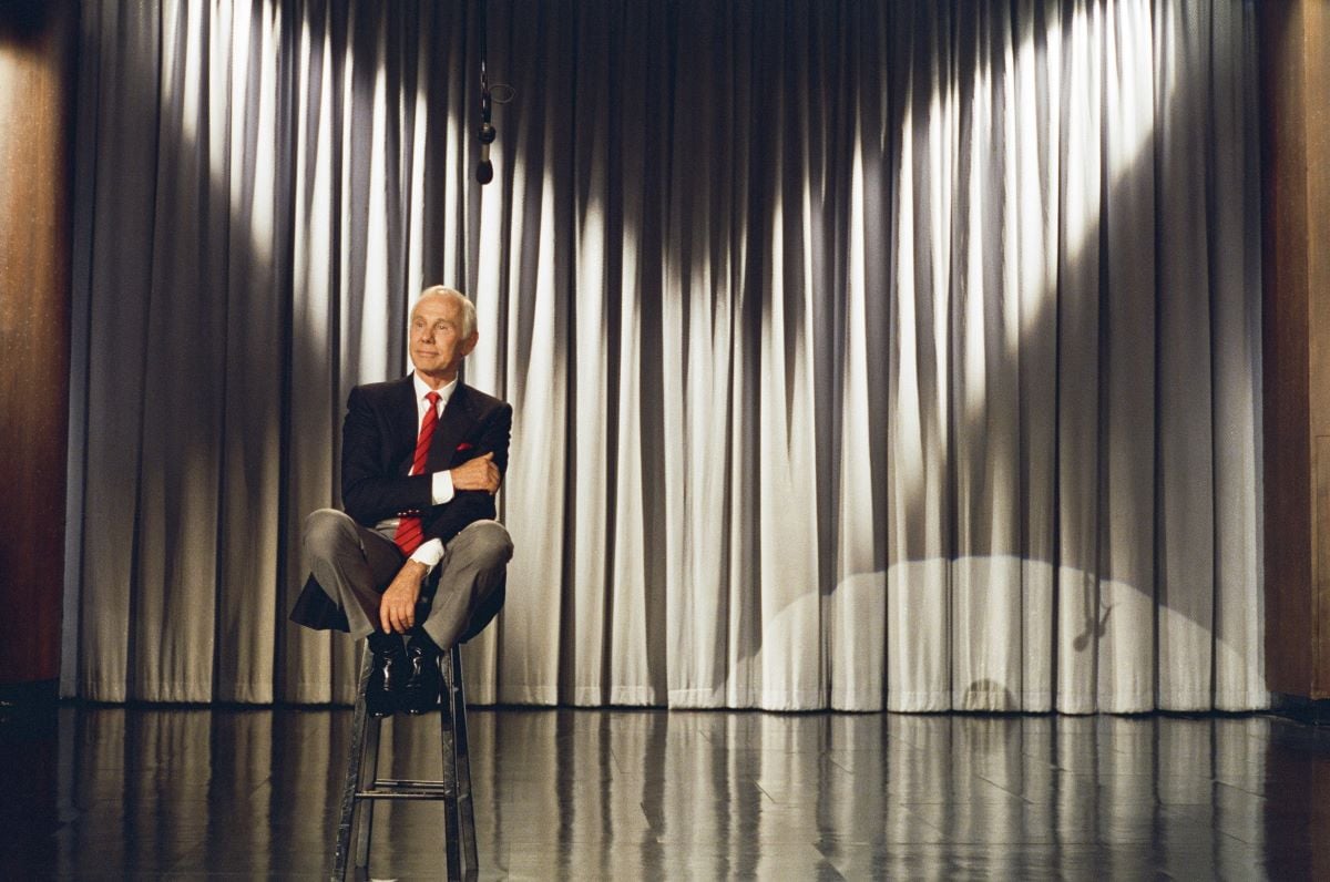 Johnny Carson seated onstage on a stool, his knees pulled up