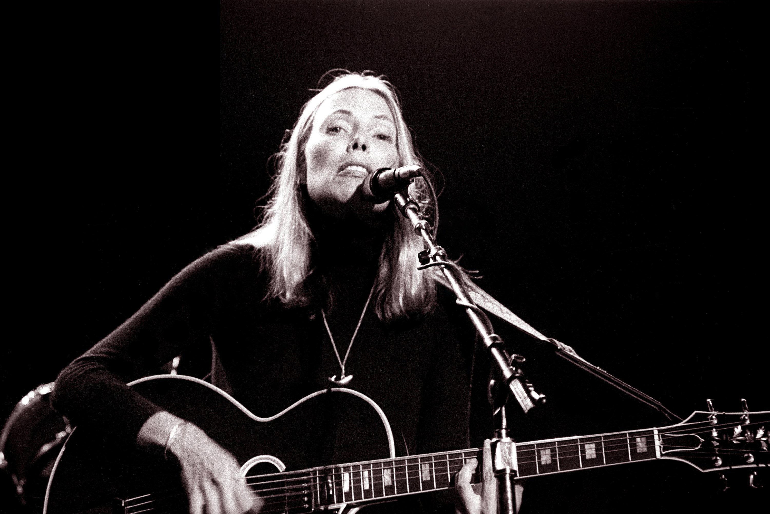 Joni Mitchell wears a black turtleneck and plays the guitar in front of a microphone.