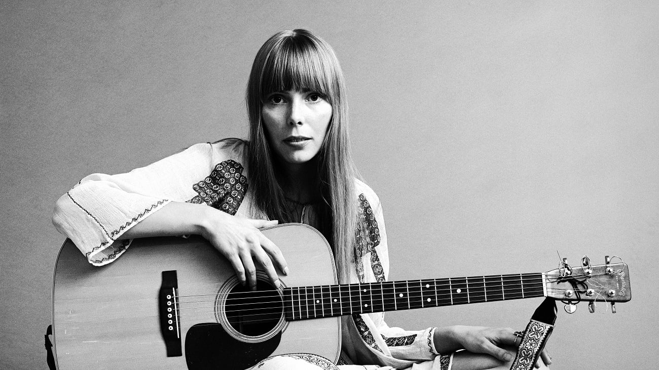 Joni Mitchell wears a white embroidered shirt and sits with a guitar.