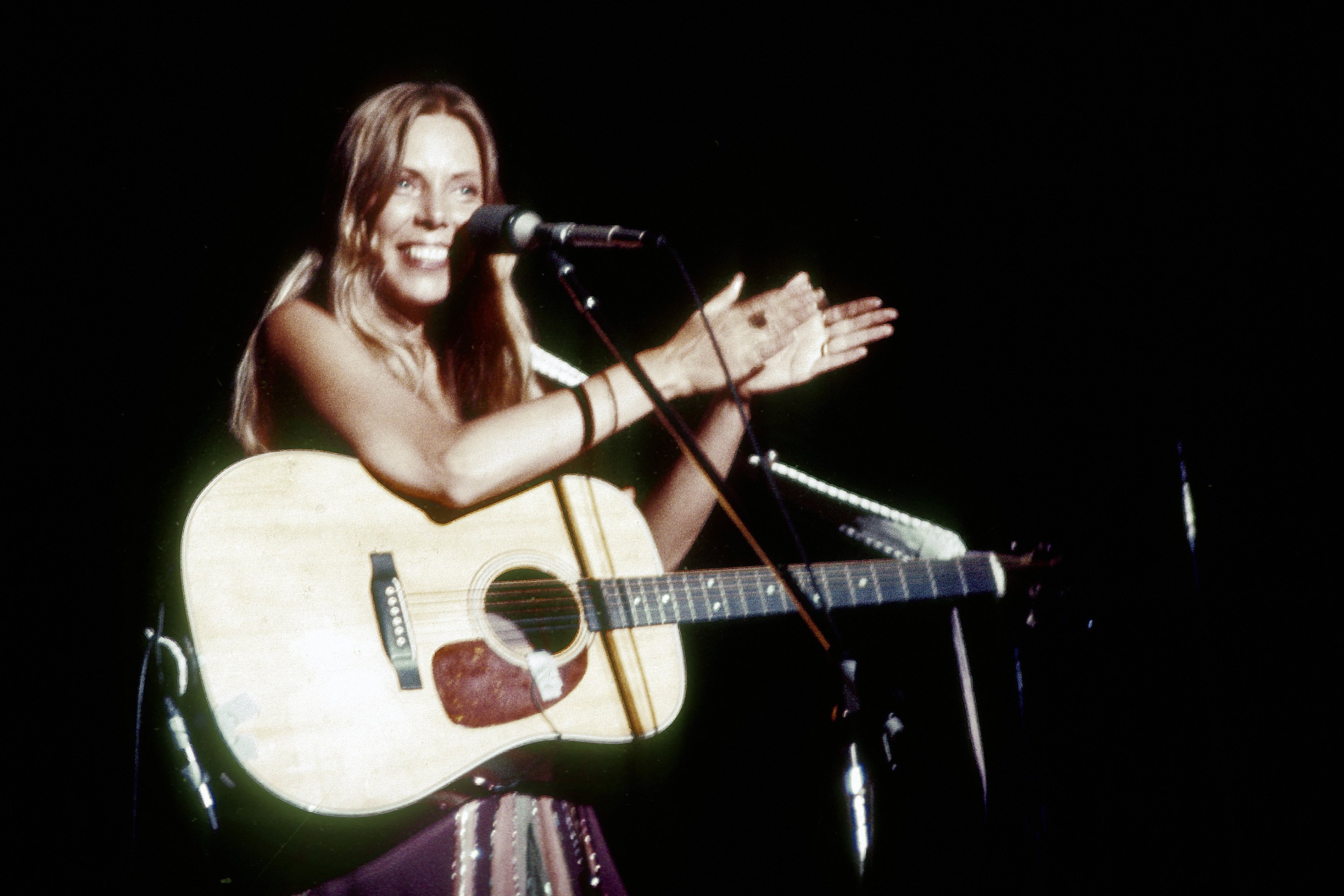 Joni Mitchell stands in front of a microphone and holds a guitar.