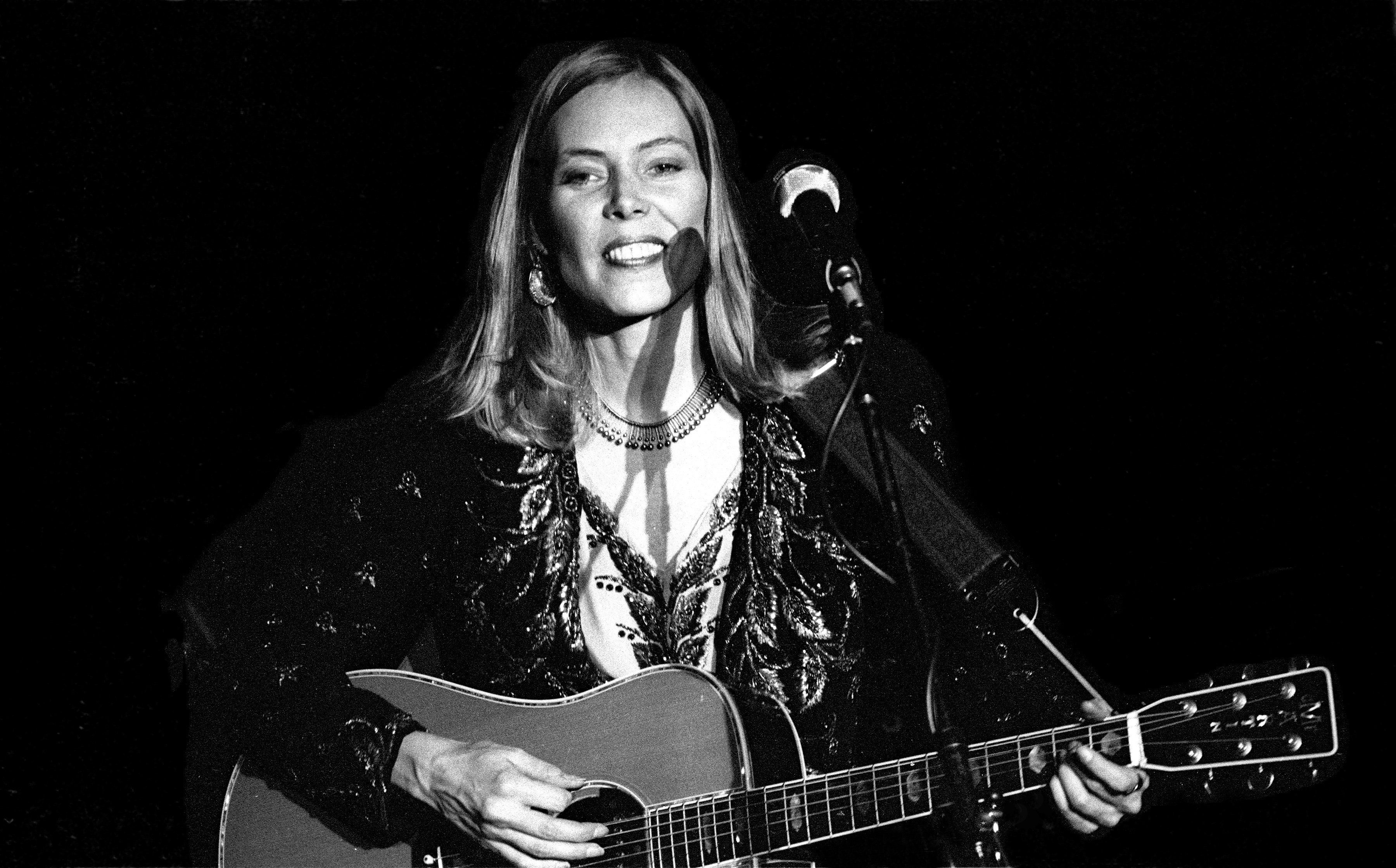 A black and white photo of Joni Mitchell wearing a black shirt and strumming a guitar. She stands in front of a microphone.