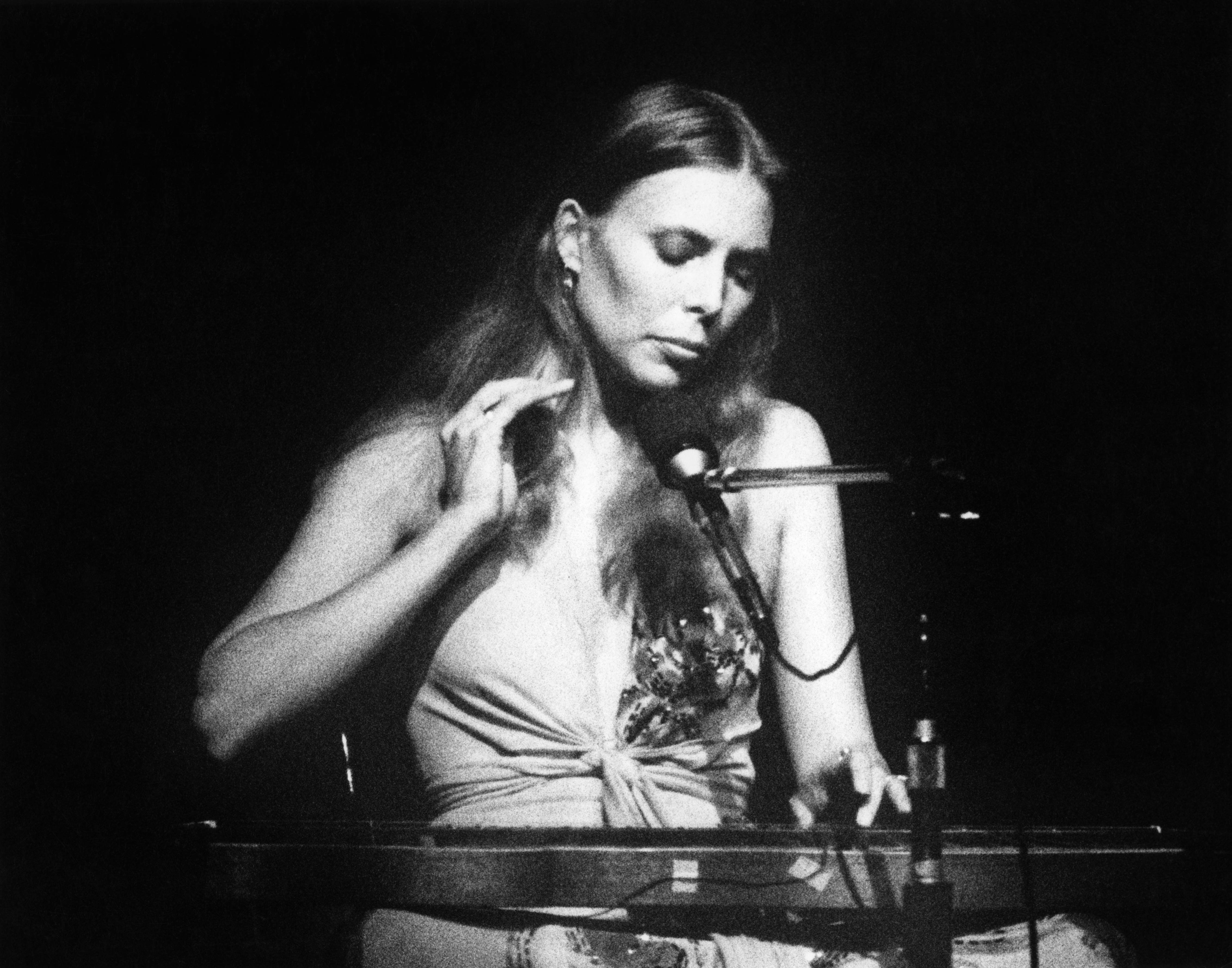 Joni Mitchell wears a white tank top and sits at a piano with a microphone.