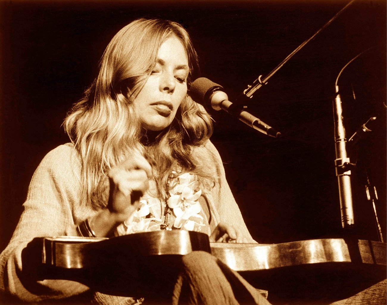 Joni Mitchell wears a sweater and a lei. She sits in front of a microphone.