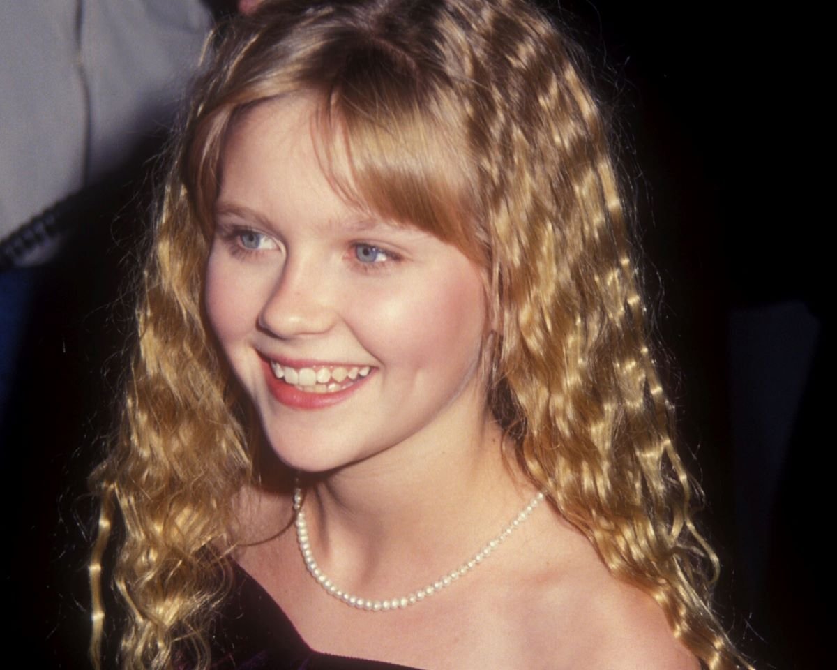 Young Kirsten Dunst with crimpy hair in an off-the-shoulder dress and pearl necklace