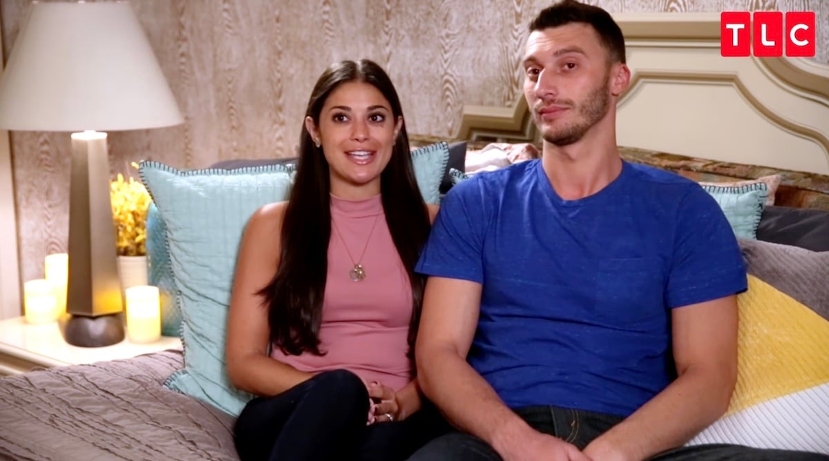 Loren and Alexei, who added to the number of 90 Day Fiancé babies this year, are seen here sitting on a couch next to one another. Loren wears a pink sleeveless top while Alexei wears a blue tshirt.