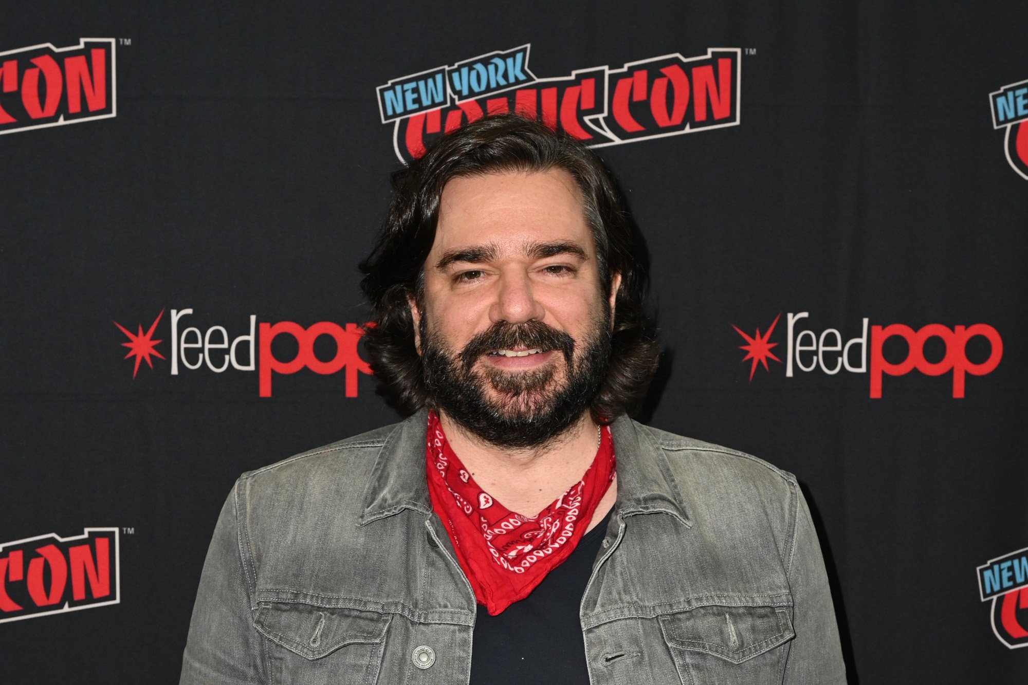'The Book of Boba Fett' and 'What We Do in the Shadows' star Matt Berry. He's wearing a denim jacket and red bandana around his neck and standing in front of a New York Comic-Con wall.