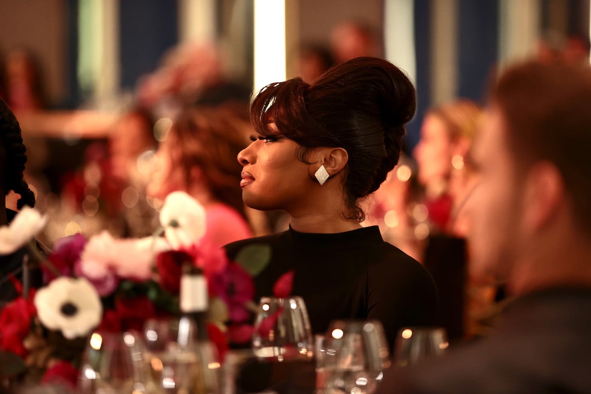 Megan Thee Stallion wearing a black top and looking to the side while seated at a table during an event.