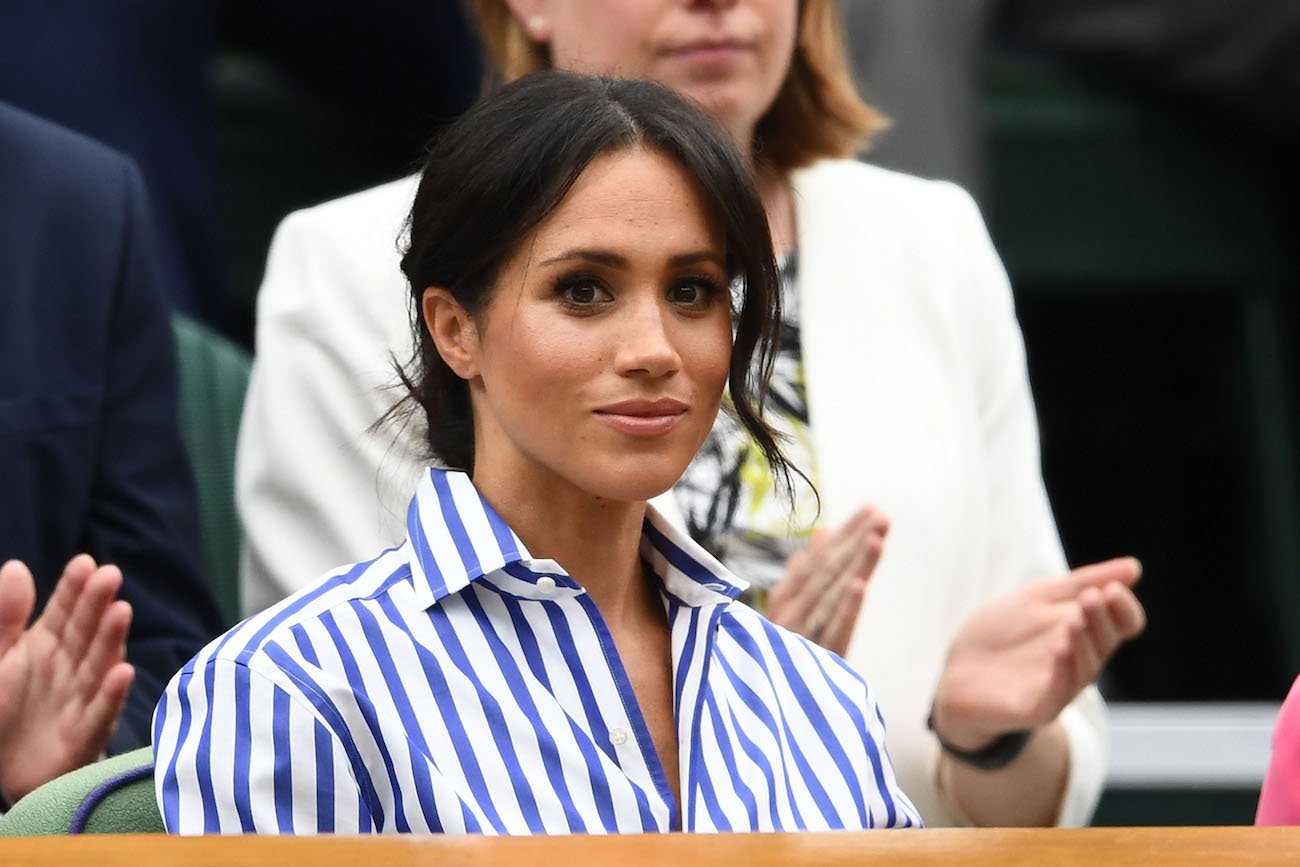 Meghan Markle looking on, wearing a blue-and-white striped shirt with her hair up
