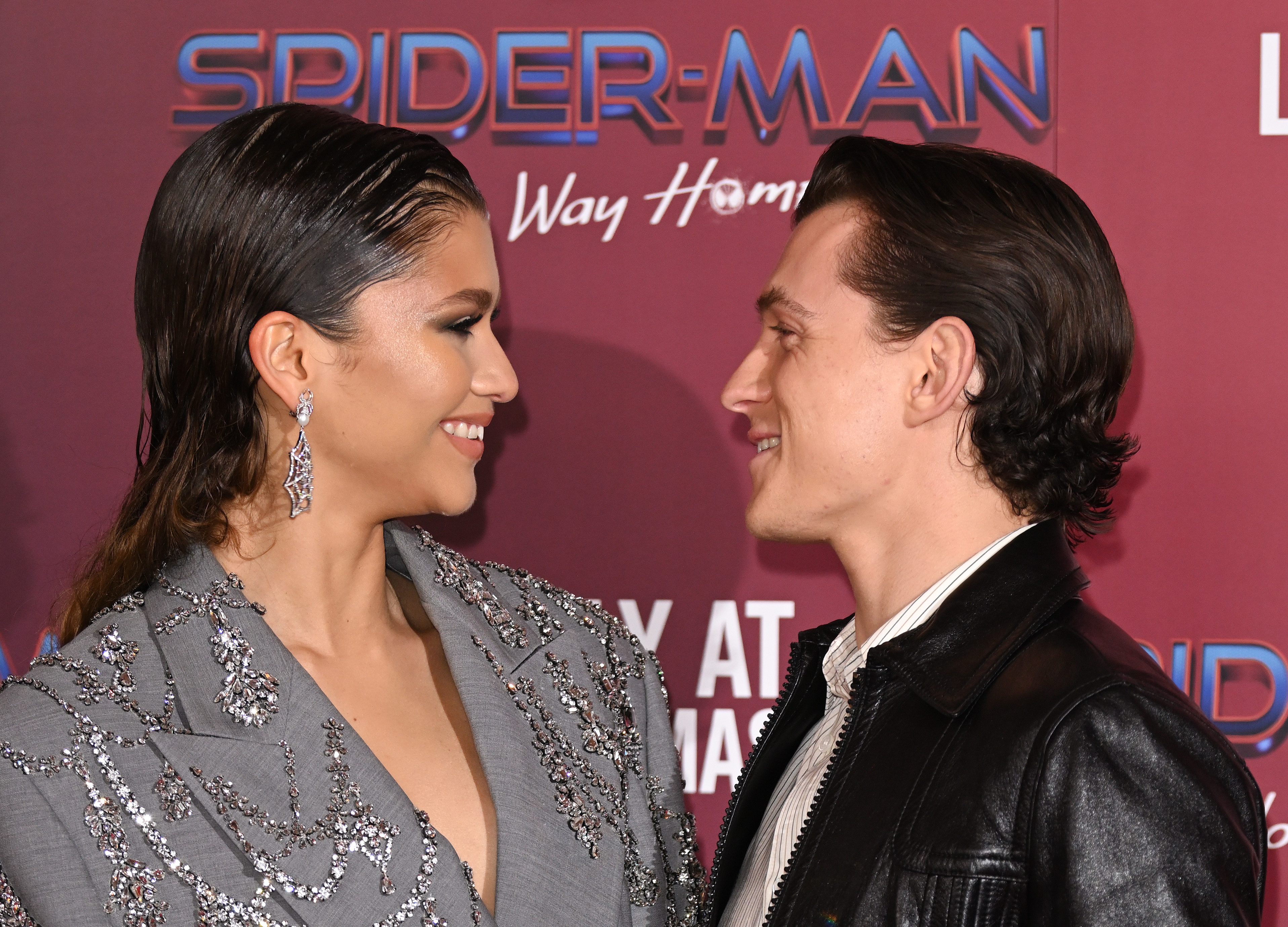 Marvel's 'Spider-Man: No Way Home' stars Zendaya and Tom Holland smile at one another as they pose for pictures. Zendaya wears a gray suit with diamond patterns on it and dangly spider web earrings. Holland wears a black leather jacket over a white striped button-up shirt.
