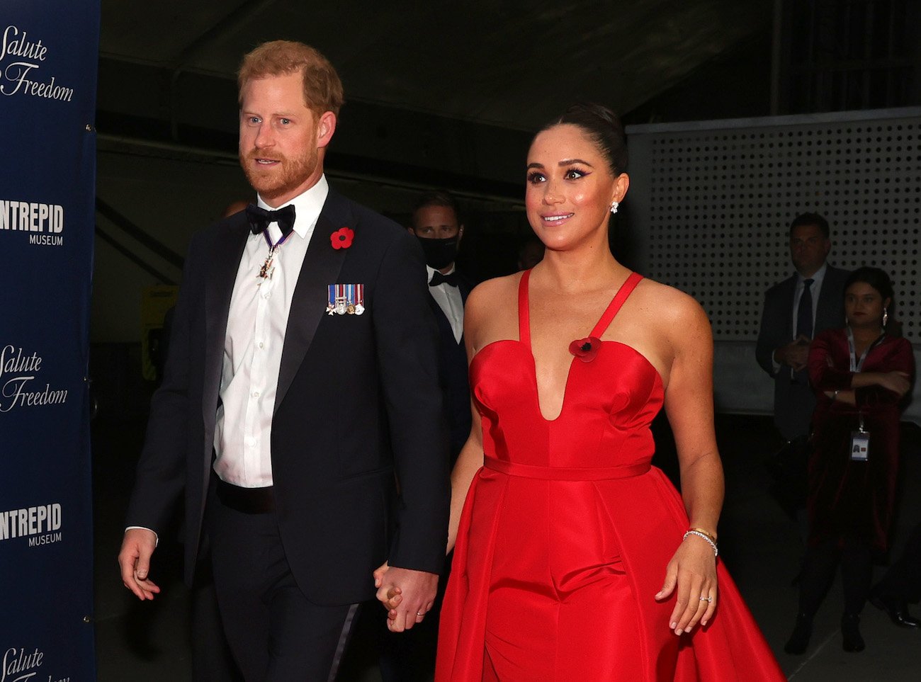 Prince Harry wears a tuxedo as he holds hands with Meghan Markle who wears a red gown