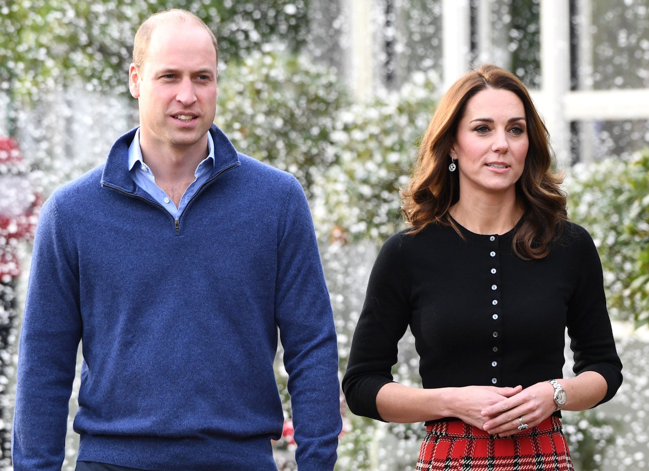 Prince William wears a blue sweater as he stands next to Kate Middleton who wears a black top and red skirt