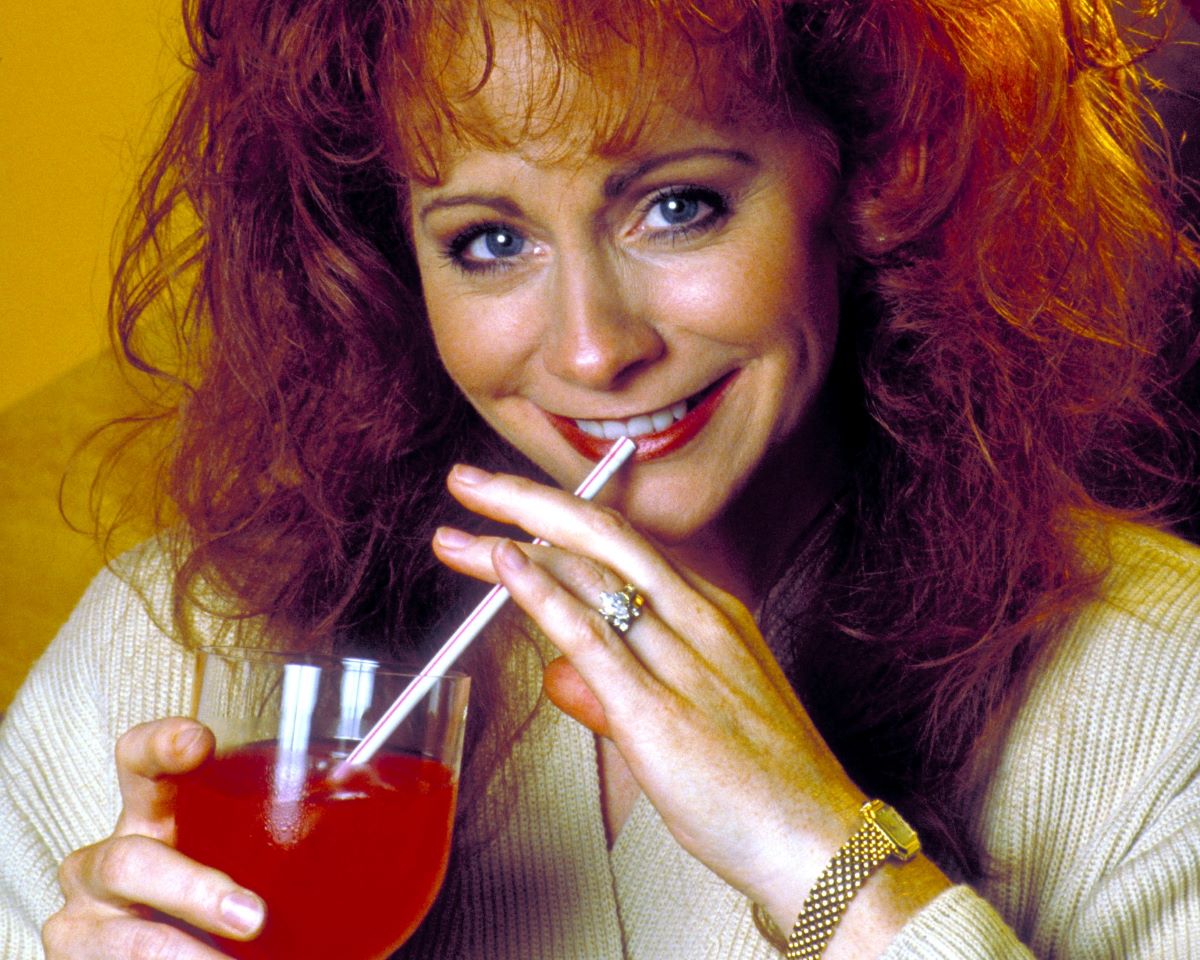 Reba McEntire places a straw from a red drink to her mouth, smiling