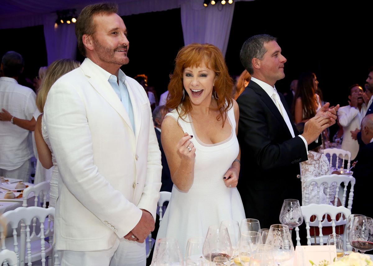 Narvel Blackstock dressed in a white suit stands next to Reba McEntire, dressed in white and smiling