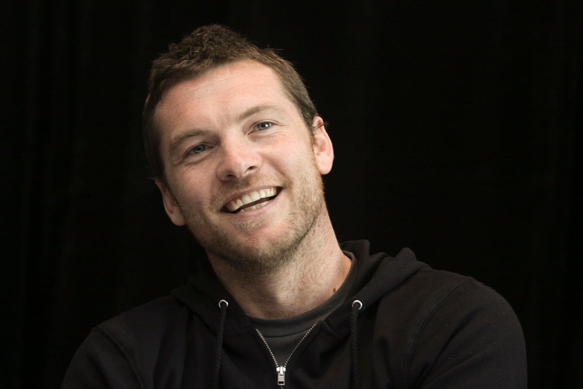 Sam Worthington smiling while wearing a black sweater.