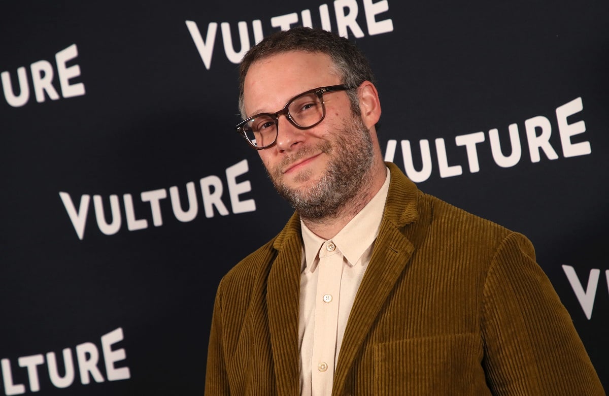 Seth Rogen smirking while wearing glasses and a brown suit.