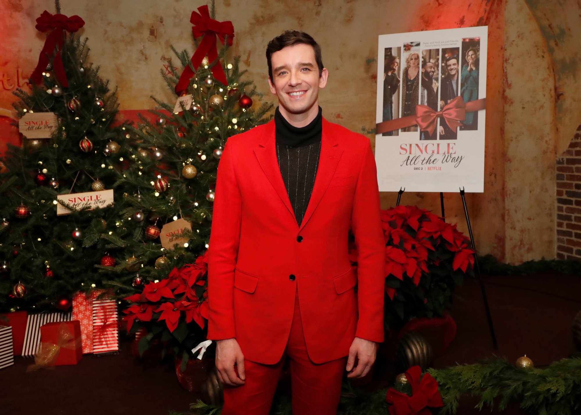 'Single All the Way' Michael Urie wearing a red suit in front of the movie's poster