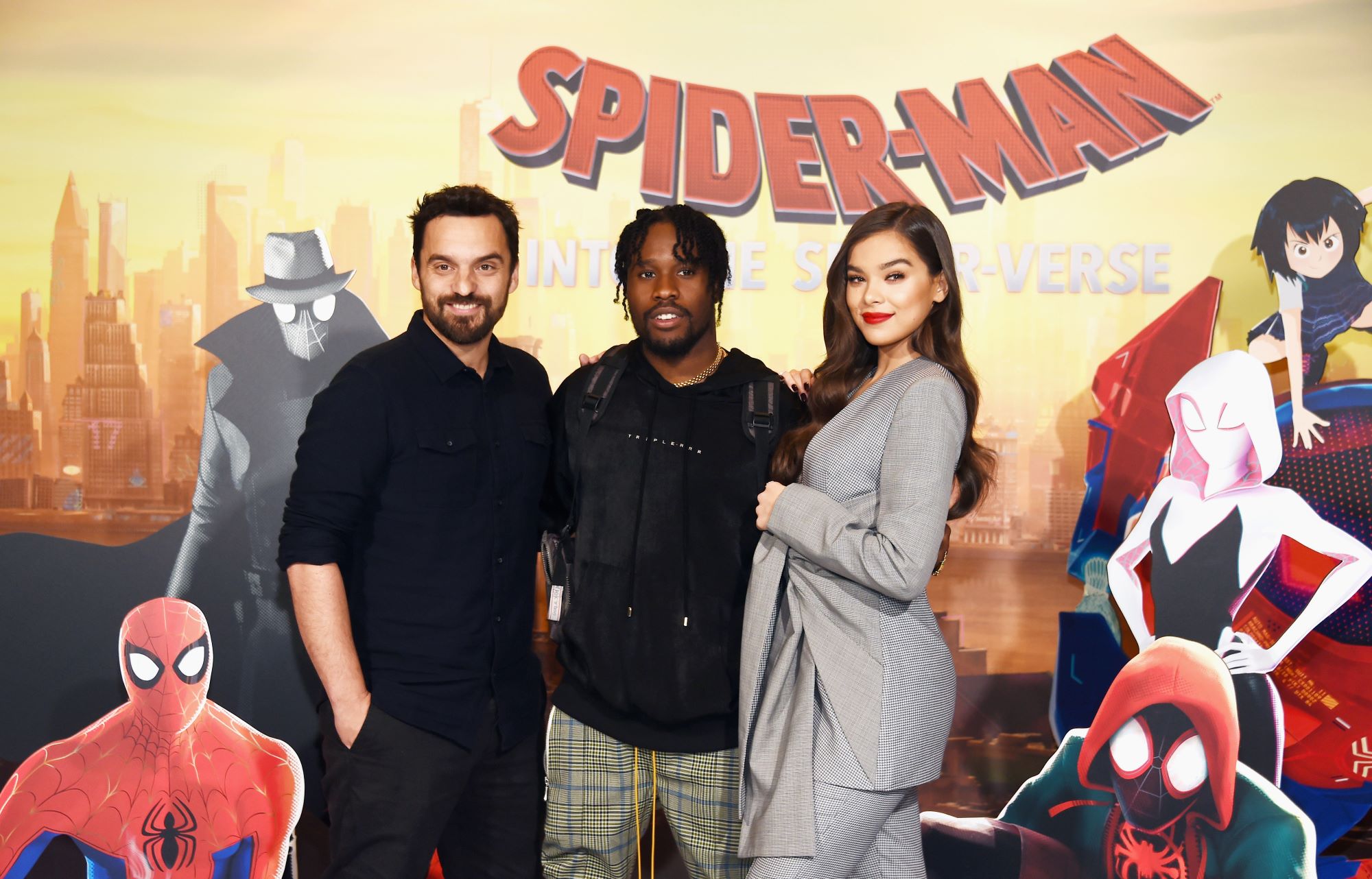 'Spider-Man: Across the Spider-Verse' stars Jake Johnson, Shameik Moore, and Hailee Steinfeld pose for a photo together. Johnson wears a black button-up long-sleeved shirt and black pants. Moore wears a black hoodie and plaid pants. Steinfeld wears a gray suit and gray pants.