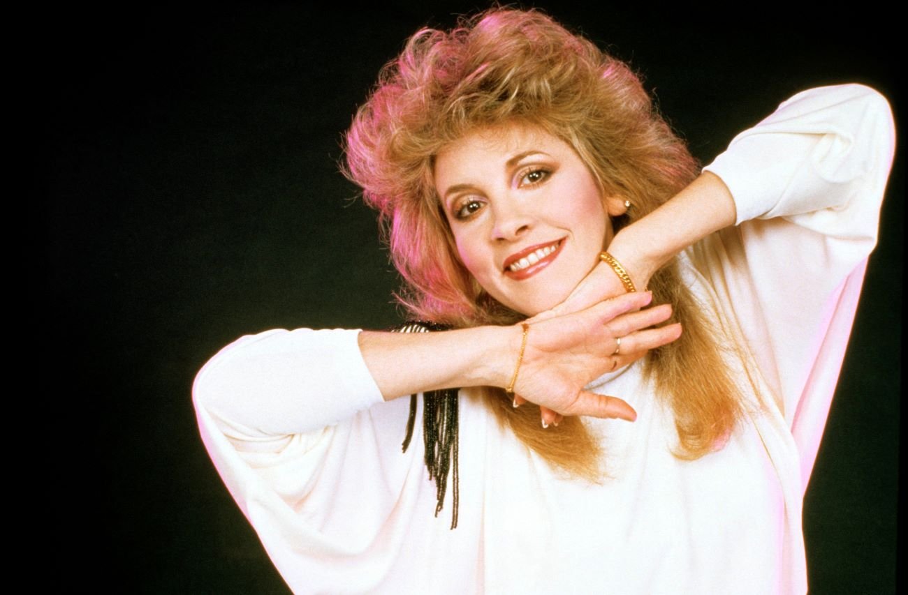 Stevie Nicks wears a white shirt and stands in front of a black background. 