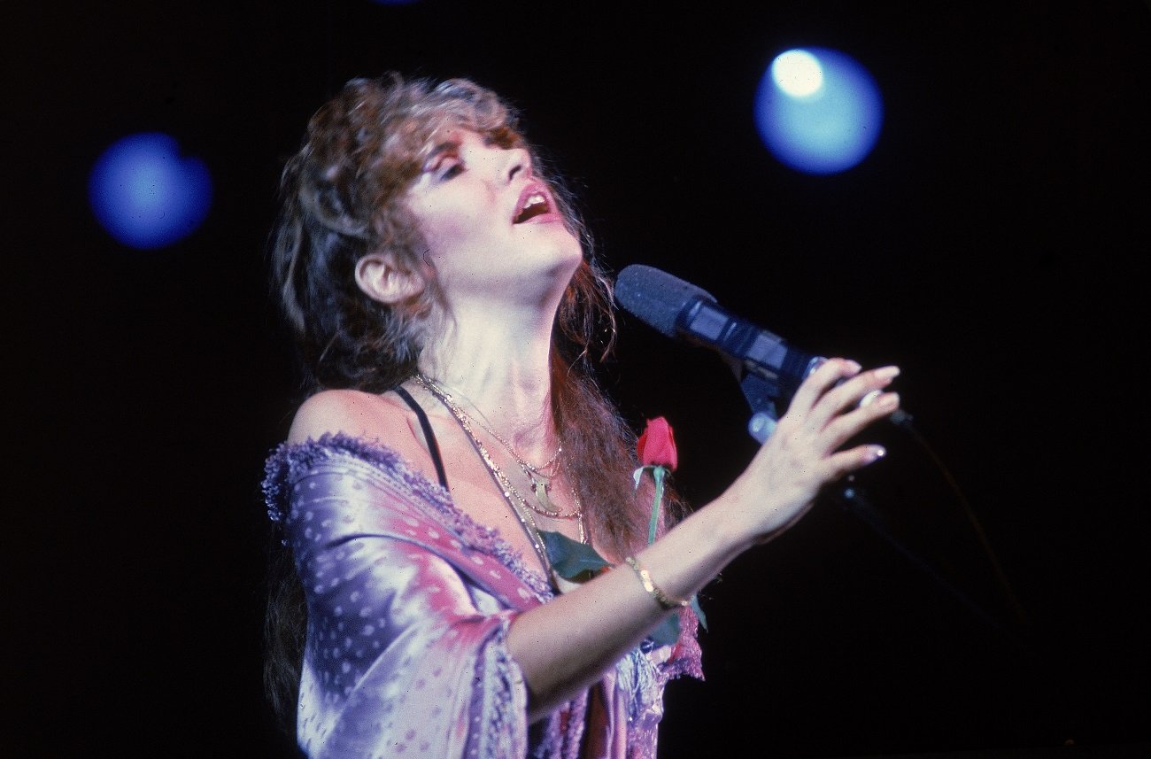 Stevie Nicks wears a pink shawl with a red rose attached and sings into a microphone. 