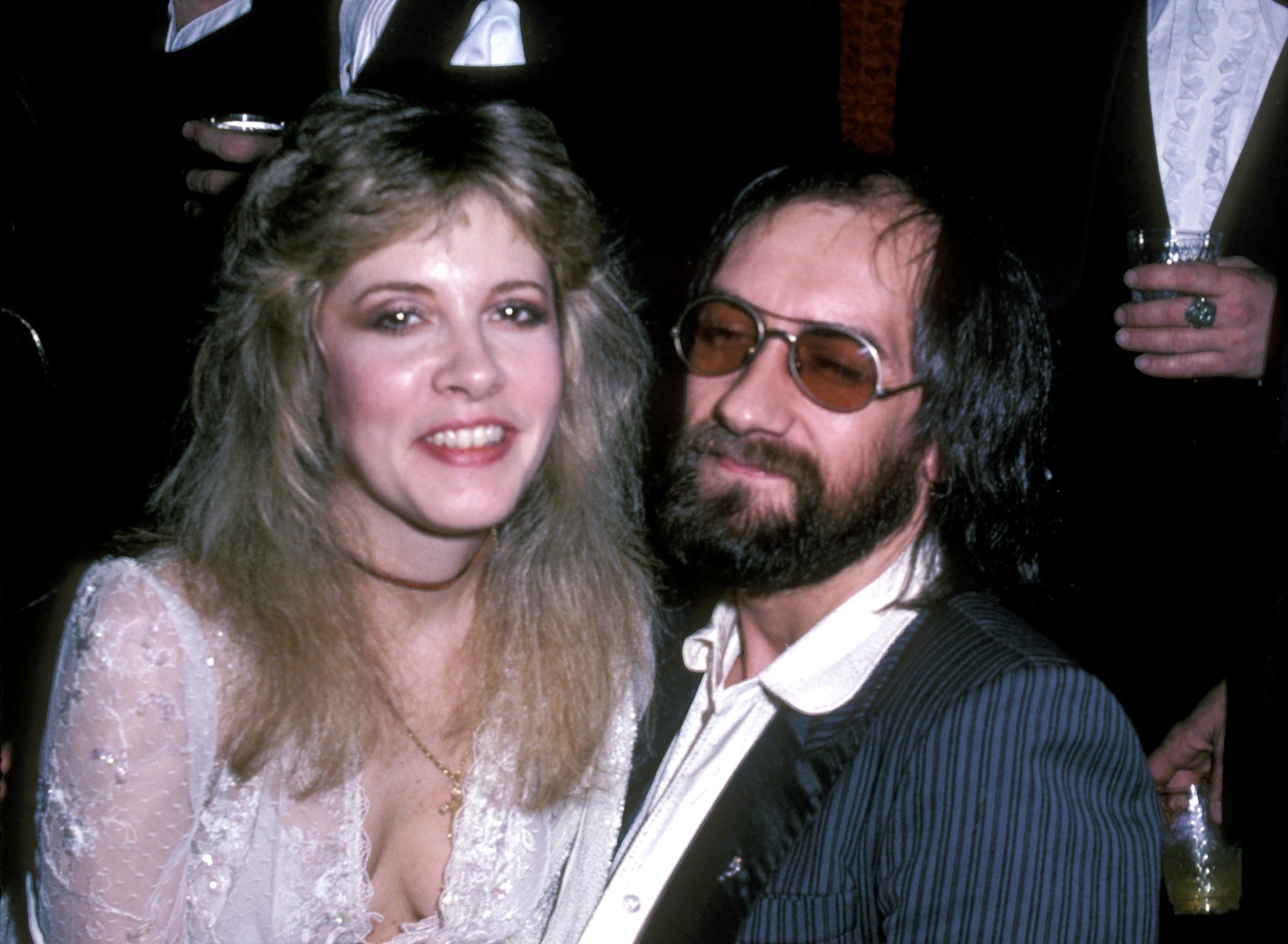 Stevie Nicks wears a white shirt and sits on Mick Fleetwood's lap. He wears a suit and sunglasses.