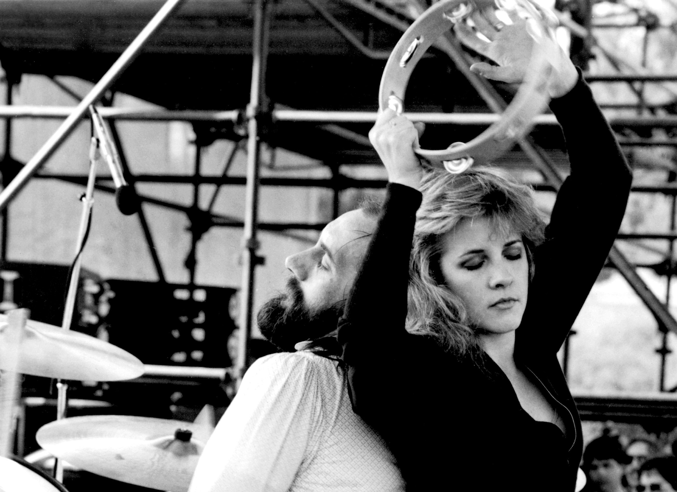 Mick Fleetwood and Stevie Nicks stand back to back while she holds a tambourine above her head.