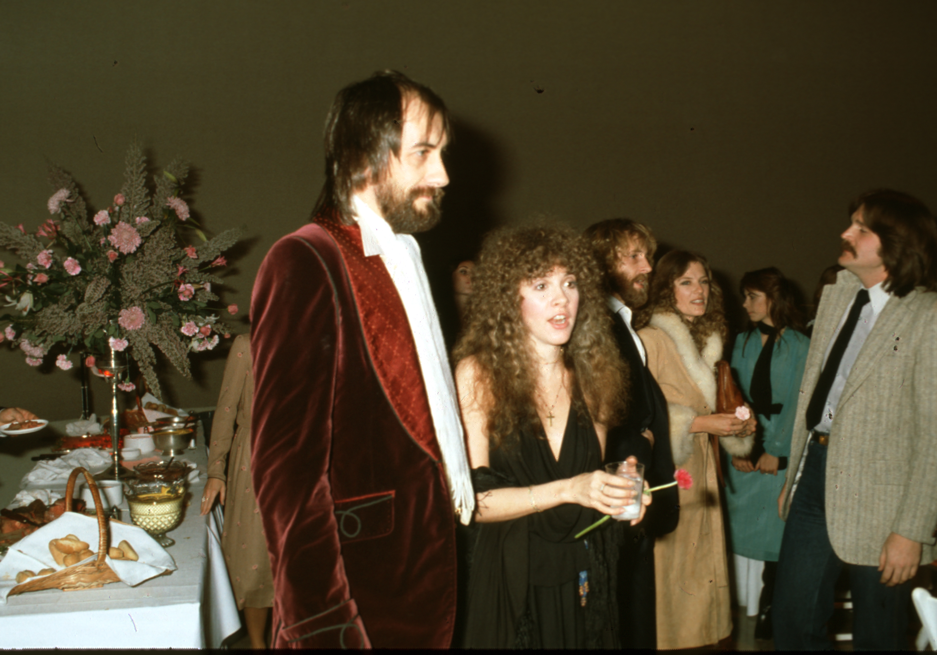 Mick Fleetwood wears a red suit and Stevie Nicks wears a black dress and holds a flower. They stand in front of a table with food.
