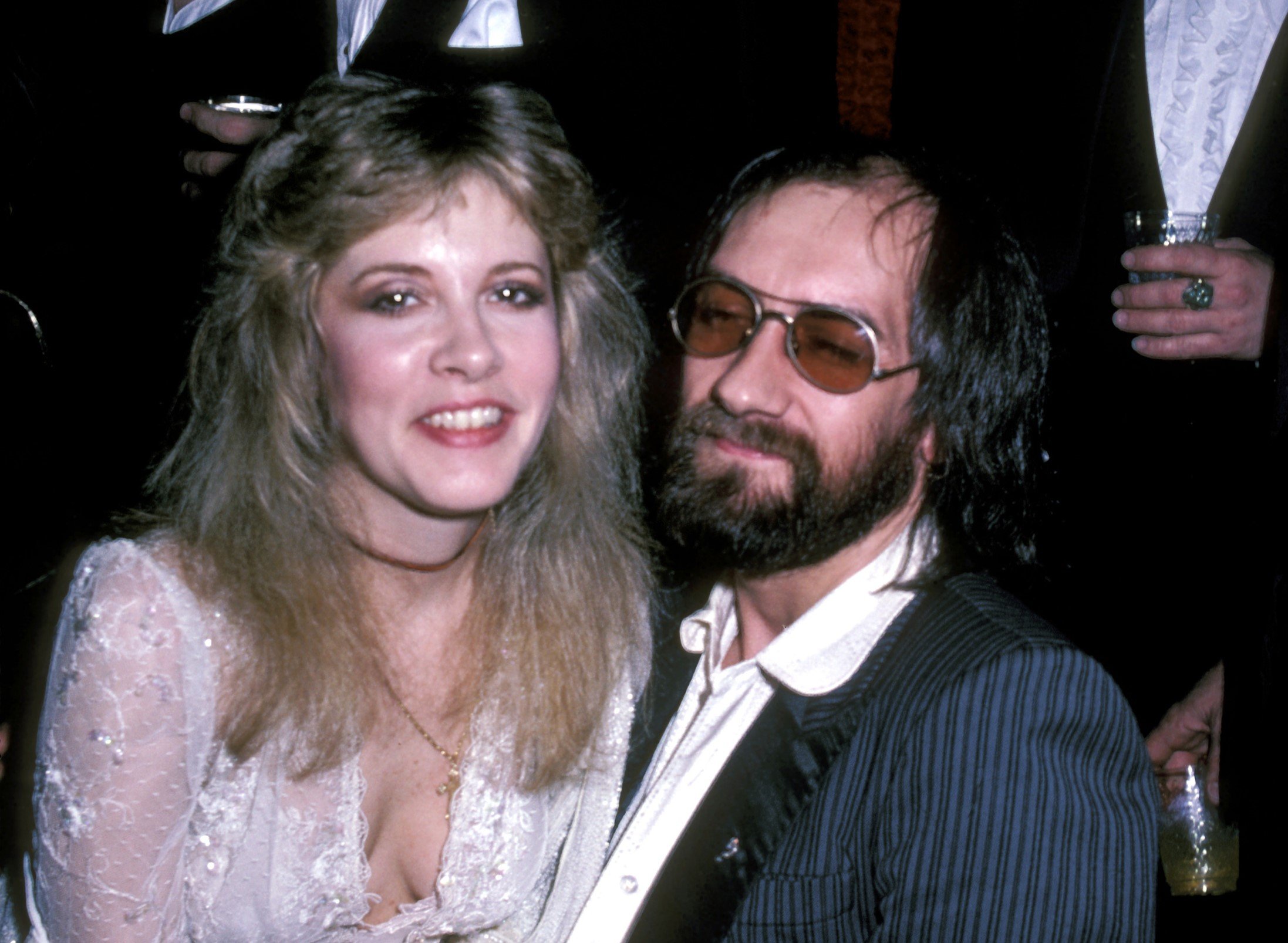 Stevie Nicks wears a white dress and sits on Mick Fleetwood's lap. He wears a blue suit jacket, white shirt, and sunglasses.