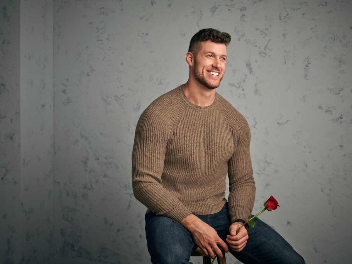 ABC's The Bachelor stars Clayton Echard. Clayton smiles wearing jeans and a sweater and holding a rose.