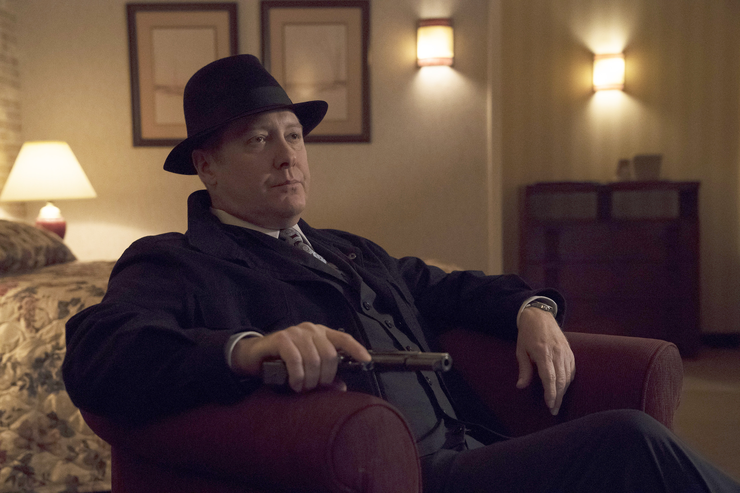 James Spader as Red in The Blacklist. Red is sitting in a chair holding a gun and wearing a hat.