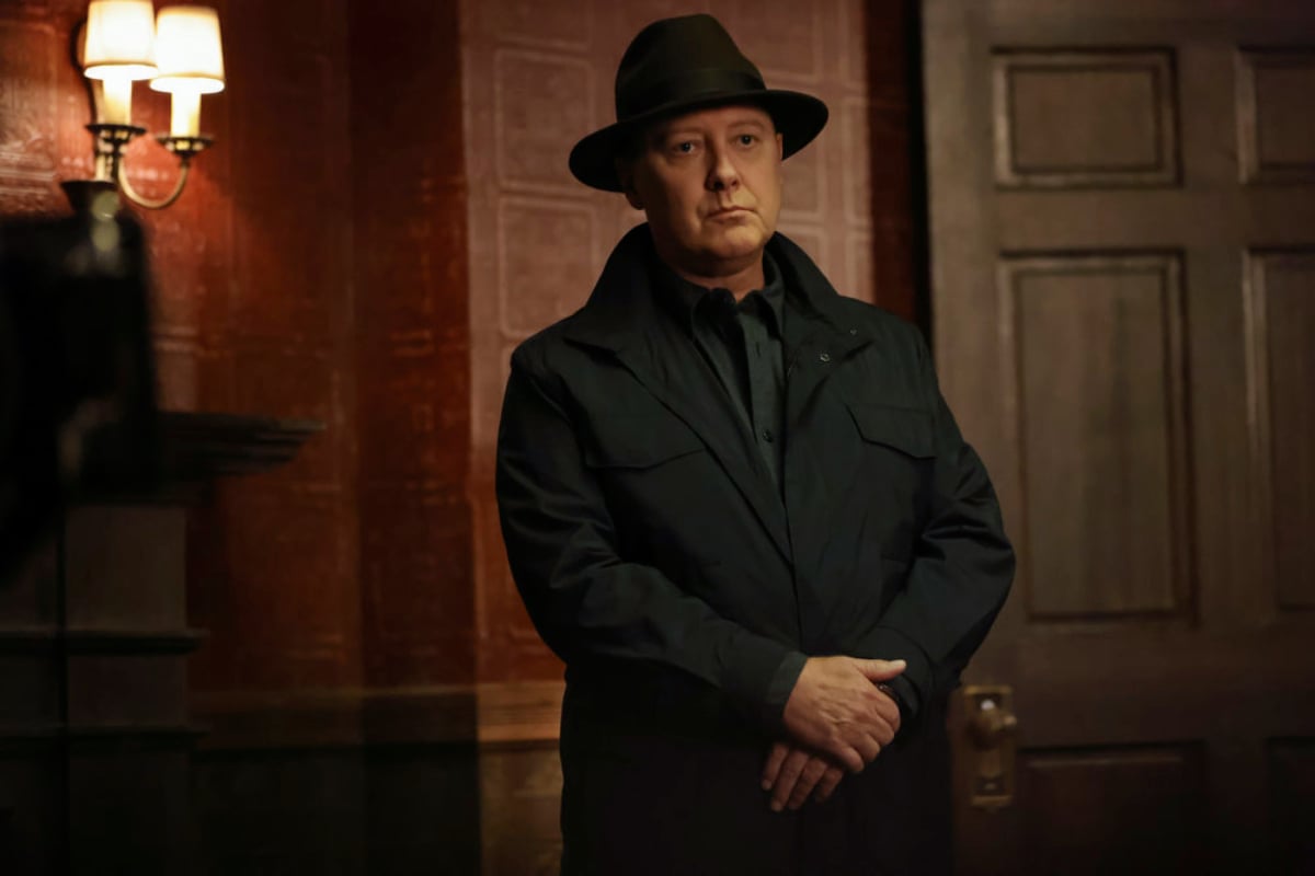 James Spader as Raymond "Red" Reddington in The Blacklist Season 9. red is wearing a hat and black coat and has his arms crossed in front of him.