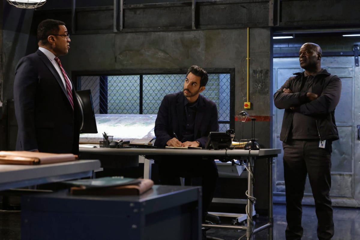 Harry Lennix as Harold Cooper, Amir Arison as Aram Mojtabai, and Hisham Tawfiq as Dembe Zuma in The Blacklist Season 9. Aram sits behind a desk. He and Dembe look at Cooper.