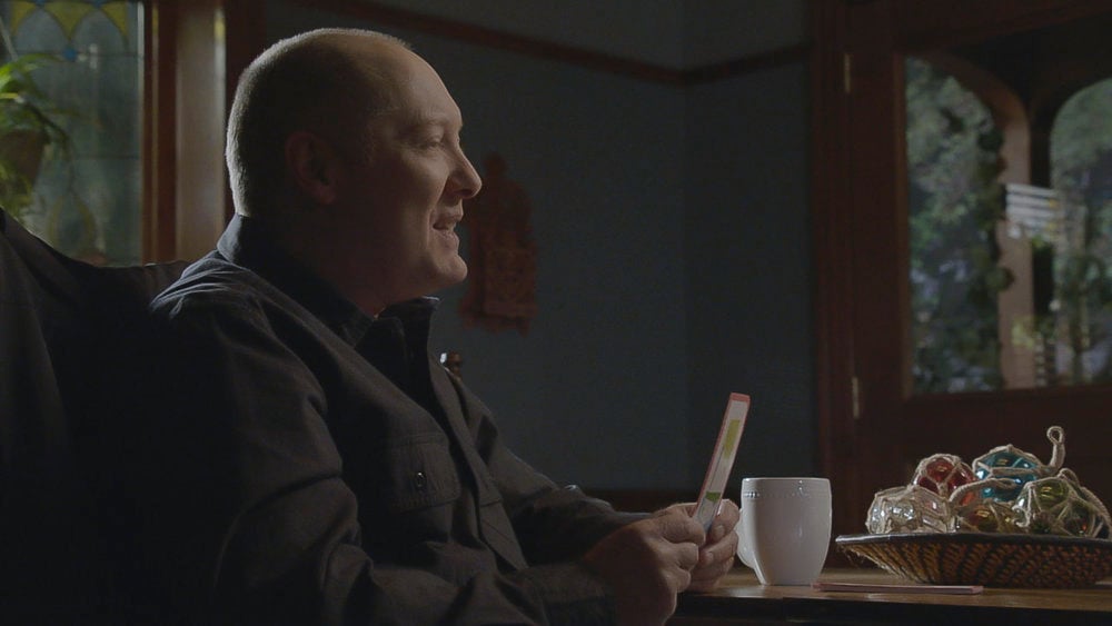 James Spader as Red in The Blacklist Season 9. Red is sitting at a table holding a card and smiling.