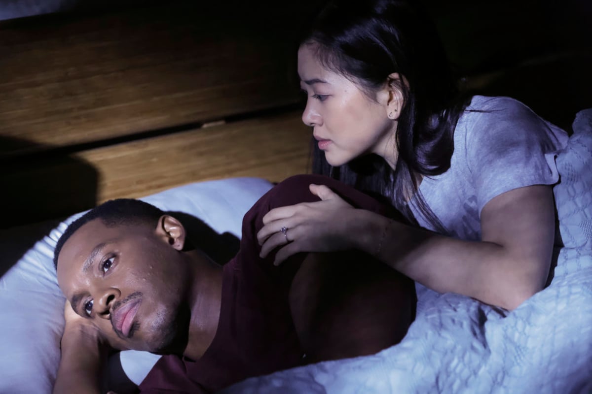 Colby Lewis as Peter Simpson and Laura Sohn as Alina Park in The Blacklist Season 9. Peter is lying in bed staring ahead and Park is looking at him.