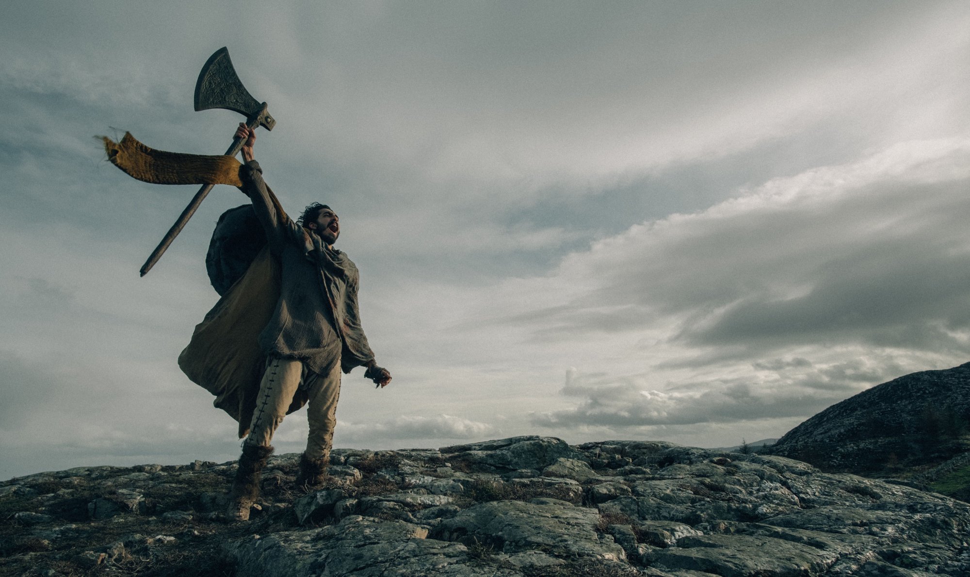 'The Green Knight' Dev Patel as Gawain holding up an axe shouting at the sky